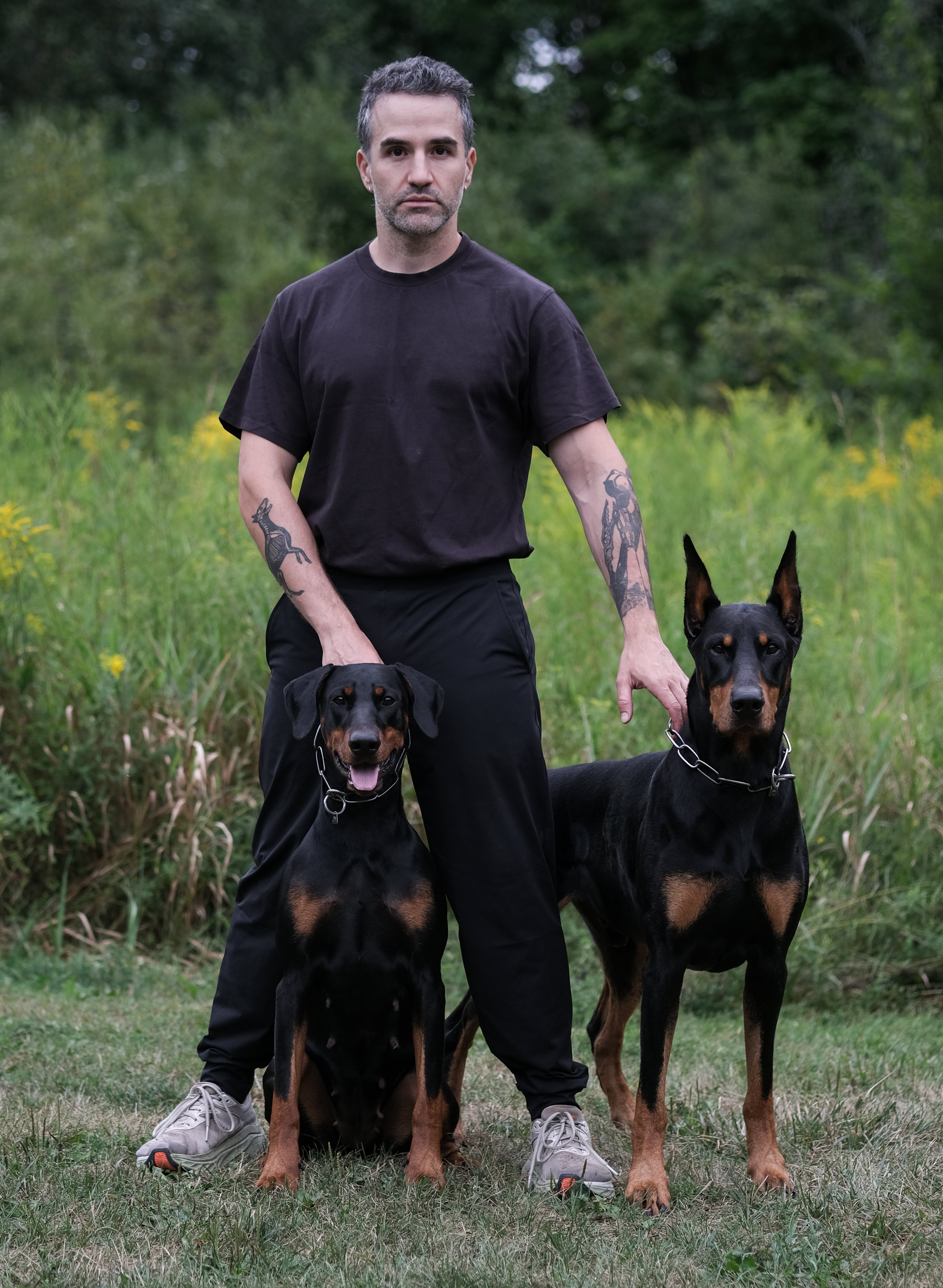 man standing with two Doberman pinschers in a grassy outdoor setting